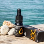 Egy üveg és egy doboz CBD Olaj 10% Full Spectrum - 1000 mg | 10 ml egy fafelületen áll, fehér és barna kövek mellett, mögötte tiszta kék vízzel. A csomagoláson sárga cseppes dizájn látható, és prémium teljes spektrumú CBD olajra utal.