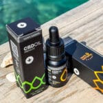 Egy üveg CBD Olaj 10% Full Spectrum - 1000 mg | 10 ml cseppentővel a fekete és zöld csomagolás mellett egy fafelületen, türkizkék vízzel és egy kagylóval a háttérben.