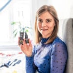 Egy vállig érő barna hajú, kék, szívmintás felsőt viselő nő ül egy ágyon és mosolyog, miközben a kezében tartja a CBD Olaj 10% Full Spectrum - 1000 mg | 10 ml terméket és annak csomagolását.