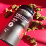 Egy piros felületen egy fekete, Pineapple Express - CBD virágok 10g | Sativa feliratú tartály áll, amelyet szétszórt szárított kannabiszrügyek vesznek körül. A címkén ananászos képek láthatók, és a termék teljes spektrumú sativa.