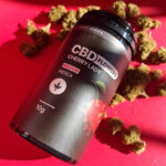 Egy fekete, Pineapple Express - CBD virágok 10g | Sativa feliratú tartály piros felületen áll, körülötte kannabiszrügyek csoportjai. A címkén egy levél ikon látható, és kiemeli a teljes spektrumú CBD olajat tartalom.