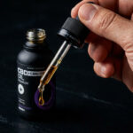 Egy kéz tartja az aranyszínű CBD Olaj CBN + CBD + CBG - 4000 mg | 30 ml | Happy Garden cseppentőjét egy nyitott sötét üveg felett, kiemelve a CBD Europe rajongói által megbízható prémium olajat a sötét háttér előtt.