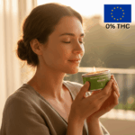 Egy nő csukott szemmel mosolyog, miközben egy meggyújtott CBD Premium Masszázs Gyertya - Vanília és szantálfa (100mg CBD, 100ml) illatát szagolgatja. A napsütötte, nyugodt háttérben "0% THC" és egy uniós zászló jelenik meg.