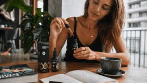 Egy nő ül az asztalnál, kezében egy cseppentővel, amelyben olaj van, és amelyet egy „CBD Oil” feliratú üveg fölé tart. Az asztalon egy csésze, egy nyitott könyv, egy jegyzetfüzet és egy termékdoboz látható. A nő nyugodtnak tűnik a világos, modern szobában.