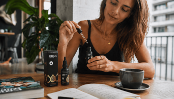 Egy nő ül az asztalnál, kezében egy cseppentővel, amelyben olaj van, és amelyet egy „CBD Oil” feliratú üveg fölé tart. Az asztalon egy csésze, egy nyitott könyv, egy jegyzetfüzet és egy termékdoboz látható. A nő nyugodtnak tűnik a világos, modern szobában.