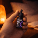 Egy kéz tartja a Happy Garden 20%-os CBD + CBN + Melatonin Olaj (10ml - 2000mg) MCT olajjal ellátott kis sötét üvegét. A meleg fényben izzó himalájai sólámpa és nyitott könyv teremt nyugodt hangulatot ennek a prémium minőségű CBD Olaj keveréknek az élvezetéhez.