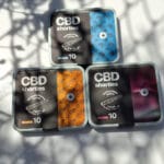 Három doboz Pineapple Express - CBD virágok 10g | Sativa feliratú konzervdoboz van elhelyezve egy fehér, leveles árnyékos felületen. Mindegyik egyedi fekete, kék vagy narancssárga dizájnnal rendelkezik, kannabisz grafikával és teljes spektrumú CBD Europe márkajelzéssel.