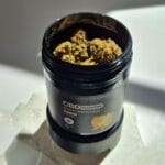 Egy fekete prémium tartály, amelyen a Pineapple Express - CBD virágok 10g | Sativa felirat szerepel, nyitva van, és a belsejében szárított kannabiszrügyek láthatók. A CBD Europeról származó tartály fehér felületen pihen, a fény lágy árnyékokat vet körülötte.
