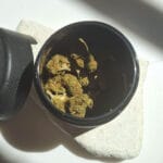 Egy nyitott fekete tartály Pineapple Express - CBD virágok 10g | Sativa egy könnyű kőfelületen ül, a fedél a közelben, a napfény árnyékot vet - megmutatva azt a minőséget, amelyről a CBD Europe ismert.