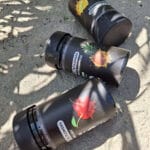 Három fekete konténer színes gyümölcsgrafikákkal (ananász, cseresznye, citrom), "Pineapple Express - CBD virágok 10g | Sativa" felirattal a homokos talajon pihen a napfényben. Prémium, teljes spektrumú kivonat. A lombozat árnyékai teszik teljessé a jelenetet.